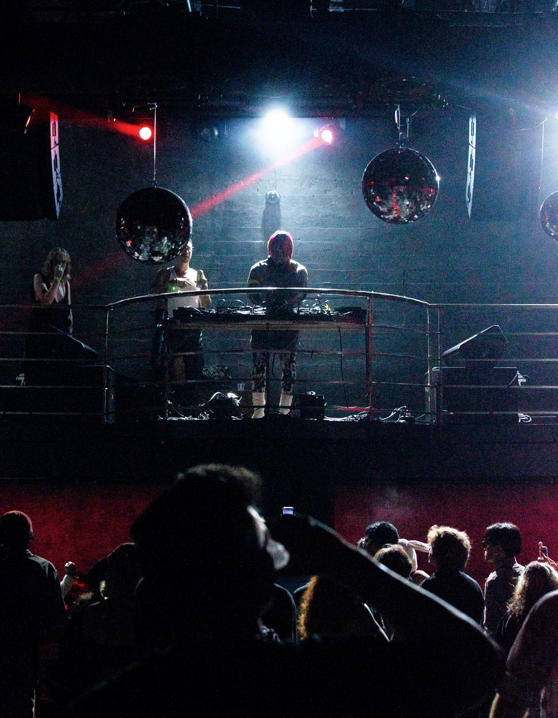 Wide shot of DJ Miss Parker on the decks in dramatic club lighting surrounded by discoballs and a dancing crowd.